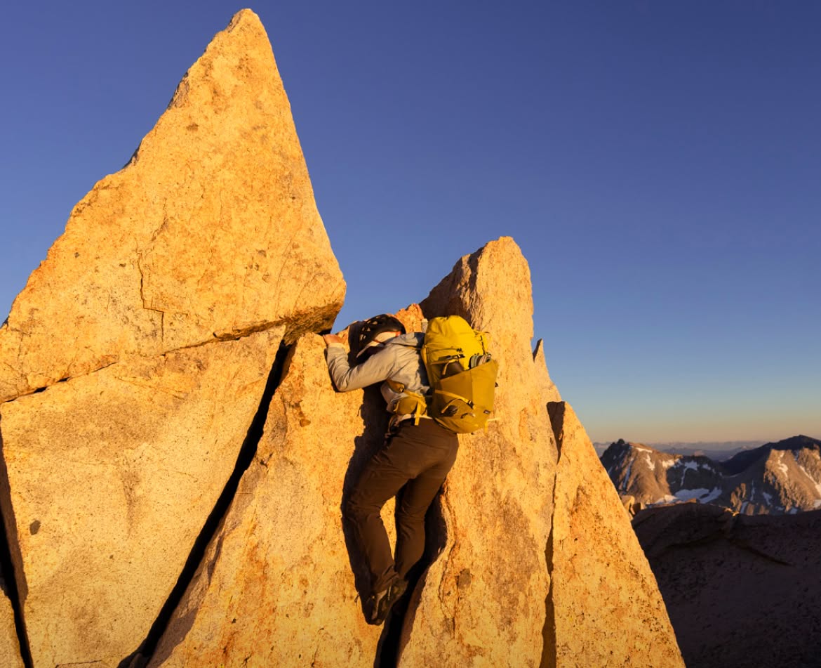 A person wearing a backpack makes a final climb to the summit of a jagged granite peak.