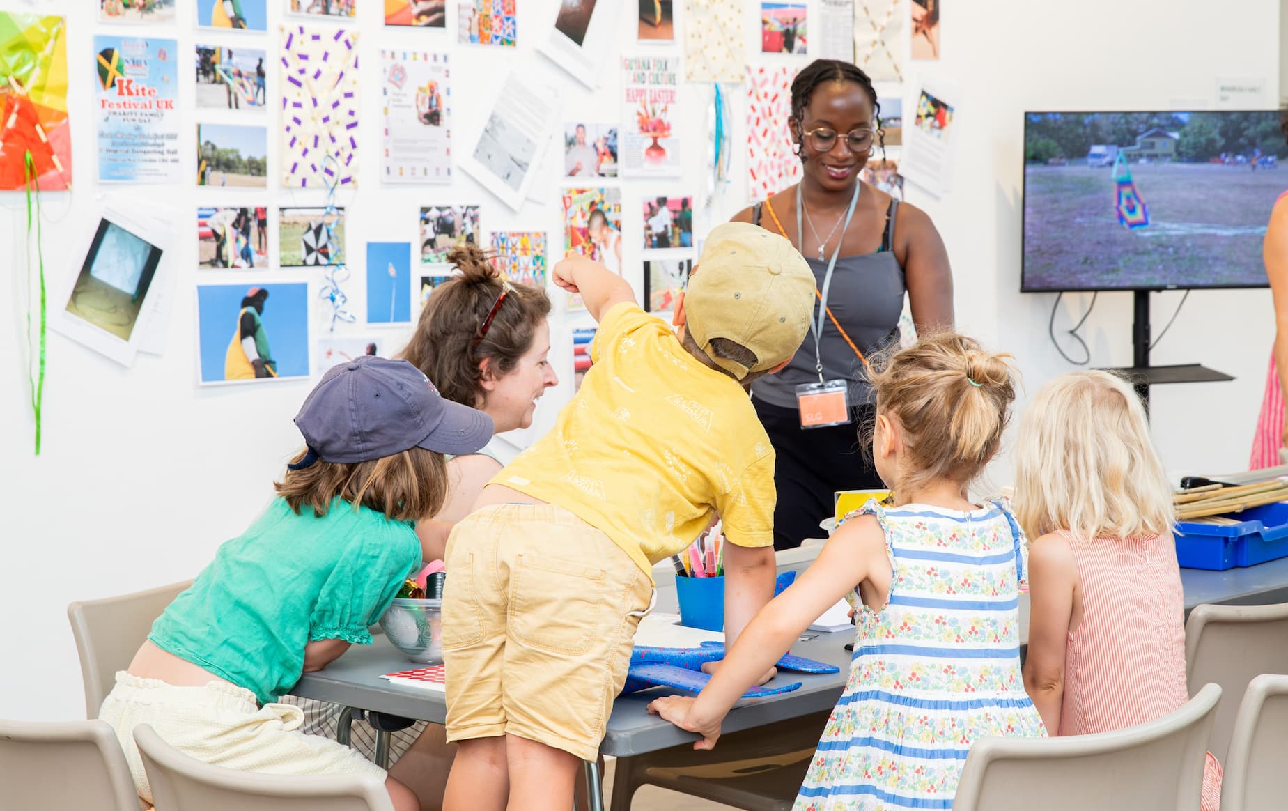 A group of five children and one adult gathered around a table during a classroom or activity session. One child in a yellow outfit stands and points upward, while the others sit on chairs facing the center of the group. The adult, wearing glasses and a lanyard, stands nearby overseeing the activity.