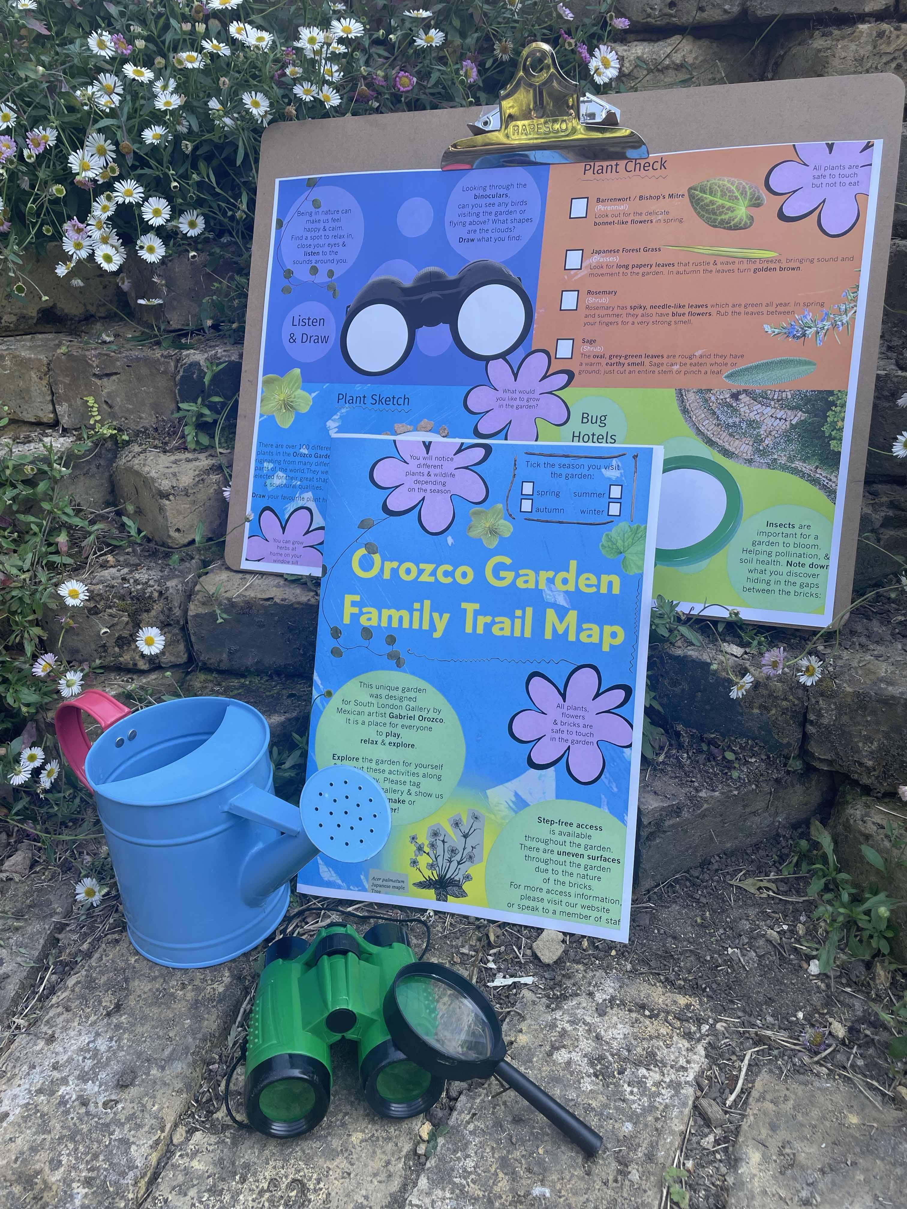 Colourful family trail map attached to flipboard against brickwork, white flowers are in the background and childrens binoculars, watering can and magnifying glass.