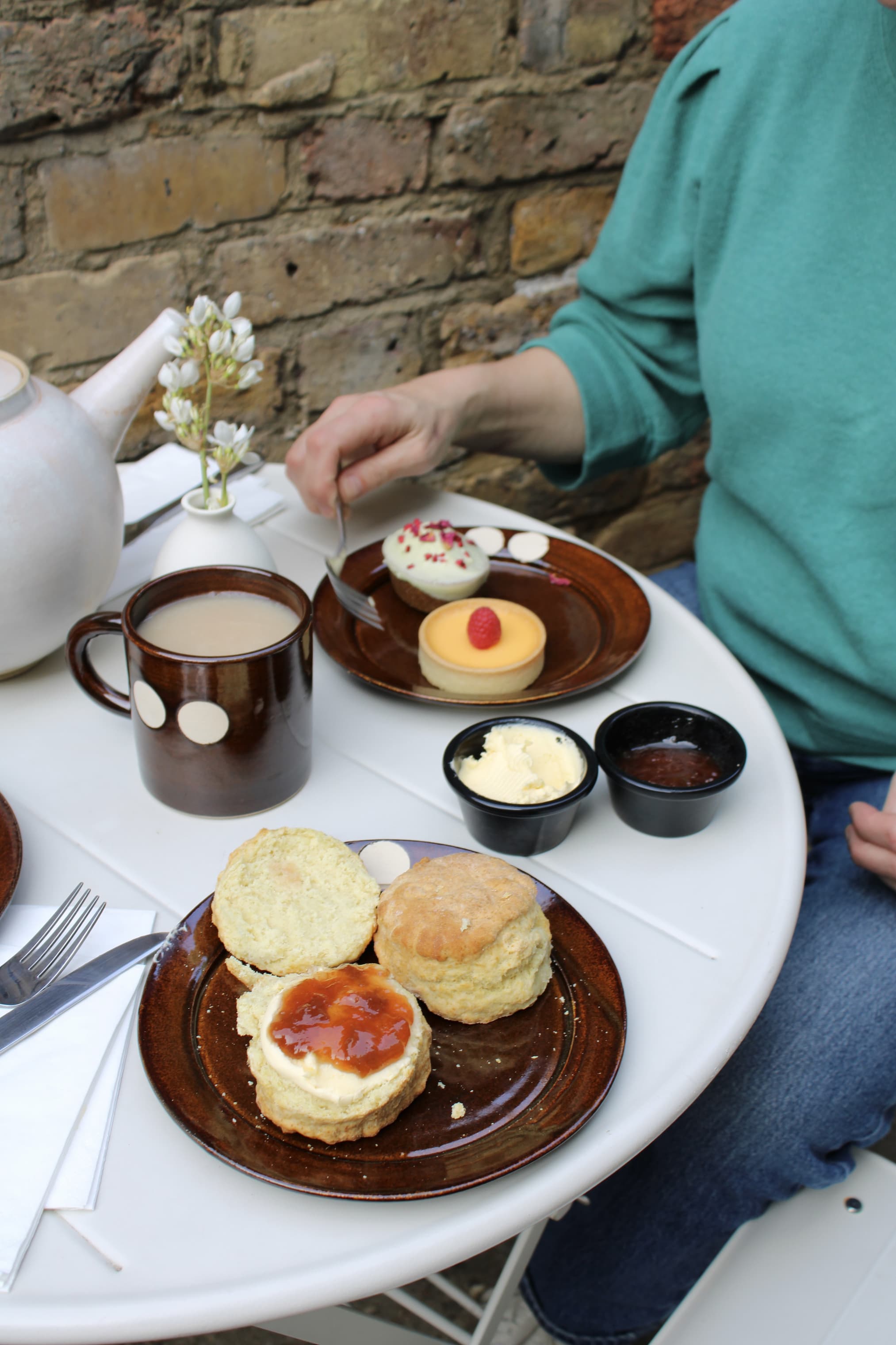 Close-up of a small café table set for afternoon tea beside a brick wall. On the table, a brown ceramic plate holds two scones, one split and topped with cream and jam. Nearby are small black ramekins of butter and jam, a cup of tea in a brown mug with cream dots, and a white teapot. A second plate holds two small desserts, including a yellow tart with a raspberry on top. A person in a green top reaches with a fork toward one dessert.