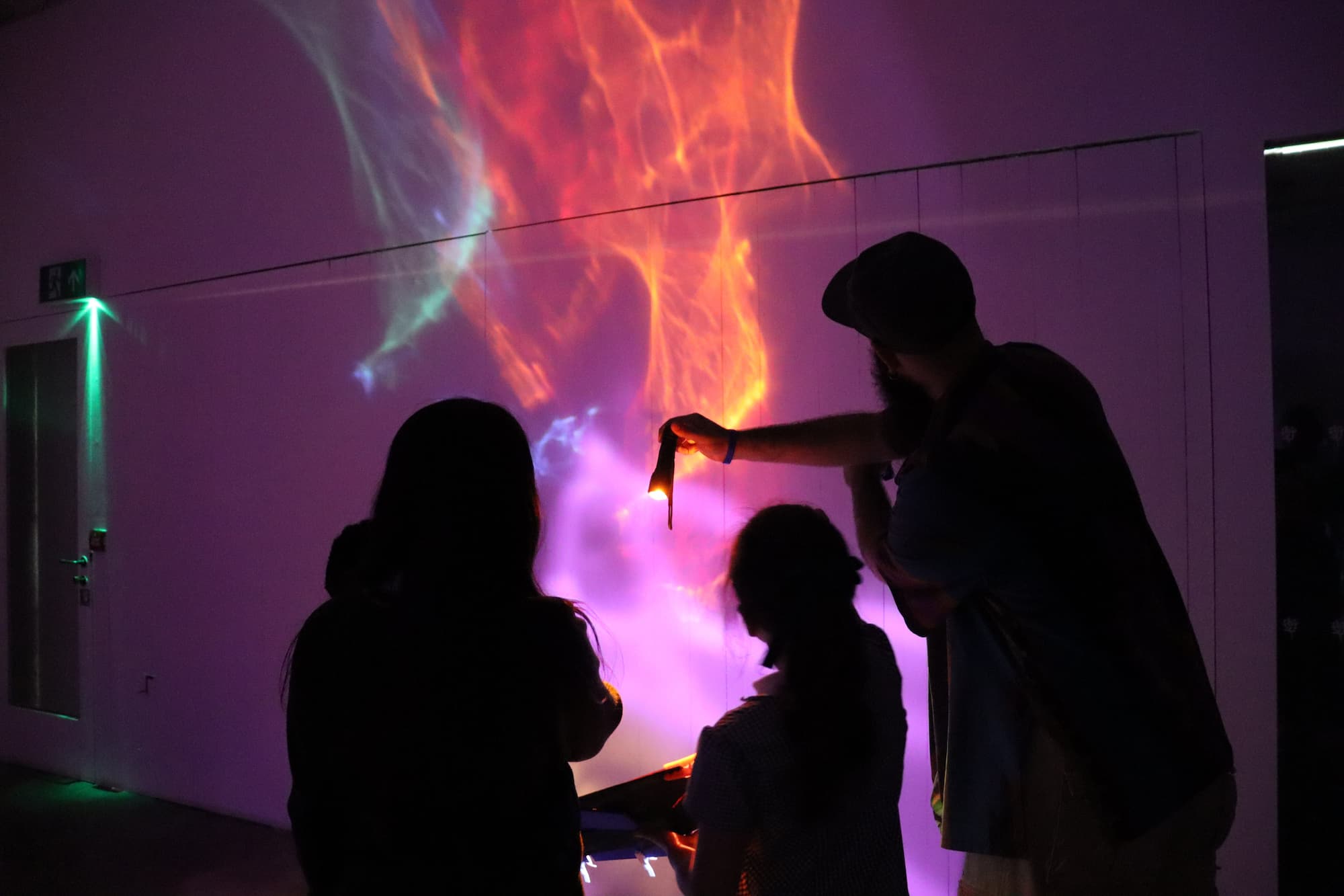 Several people stand in a dark room facing a wall illuminated by vivid projected light patterns. One person holds up a small light source, interacting with swirling pink, orange, purple, and blue visuals that resemble flames or colored smoke. The participants appear in silhouette against the bright display, creating the impression of an interactive light or digital art installation. A doorway with a green-lit exit sign is visible on the left side of the room.