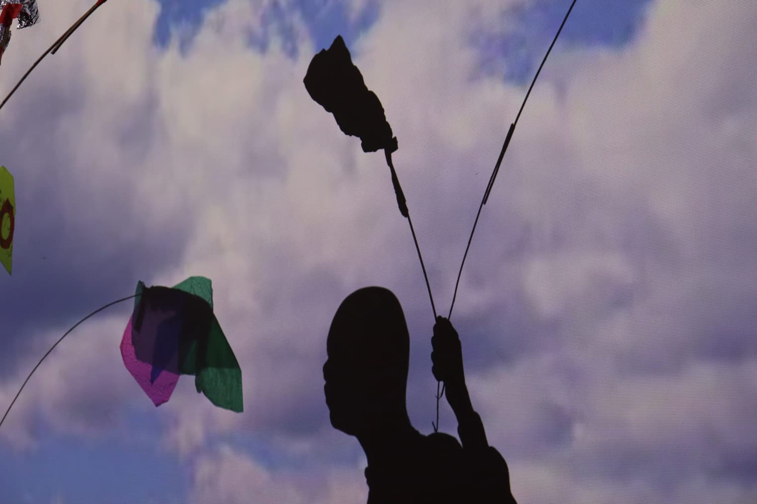 A silhouette of a person holding up a kite-like sculpture that is attached to wire. On a wall is a projected images of clouds in the sky.