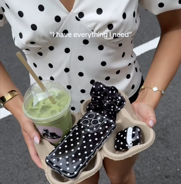 Woman in a white polka dot top holds a coffee cup holder with a green matcha, a polka dot phone case, and matching polka dot accessories.