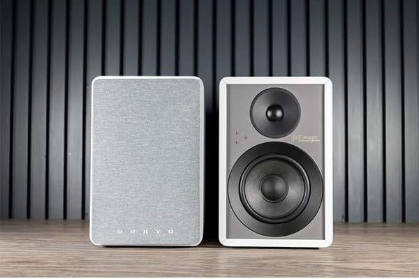 Two Onkyo GX-30ARC Powered Speakers. One showing the grille panel, the other without a grille panel. On a wooden-grain counter, in front of a black wall.