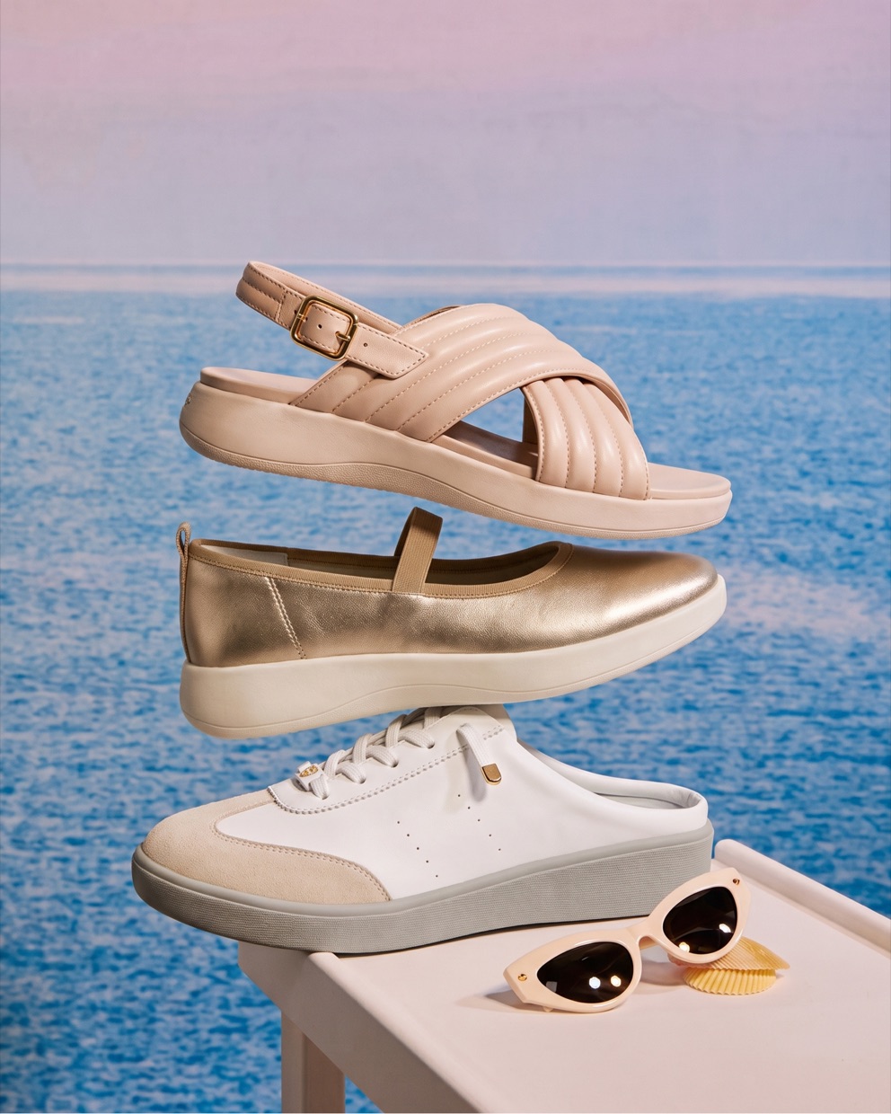 Three Sunnystep styles — a nude cross-strap sandal, a gold Mary Jane and a white sneaker — by the sea at sunset.