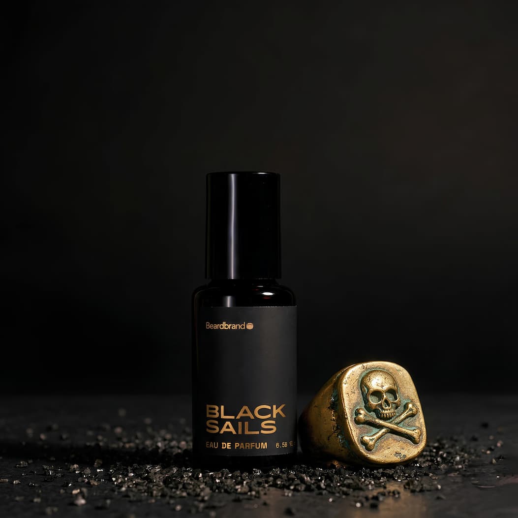 A bottle of Black Sails Cologne on a faded black background with a aged skull and cross bones ring next to it. A bottle of Black Sails Cologne on a faded black background with a aged skull and cross bones ring next to it.