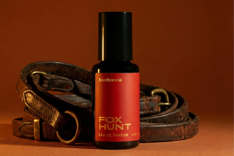 A bottle of Fox Hunt cologne on a orange backdrop with leather horse reins behind the bottle
