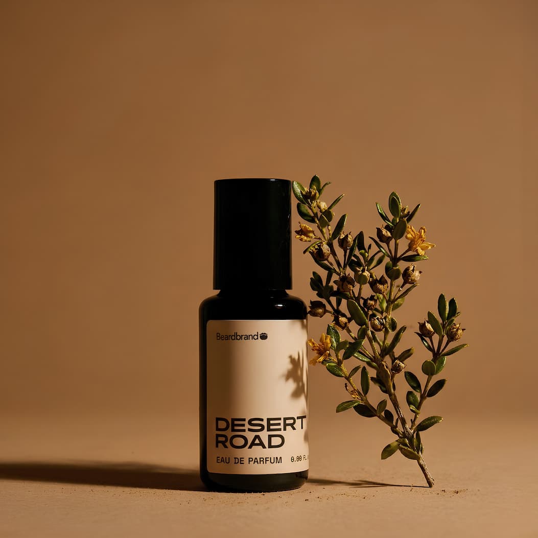 A bottle of Desert Road Cologne on a dusty tan backdrop with a single sprig of creosote. 